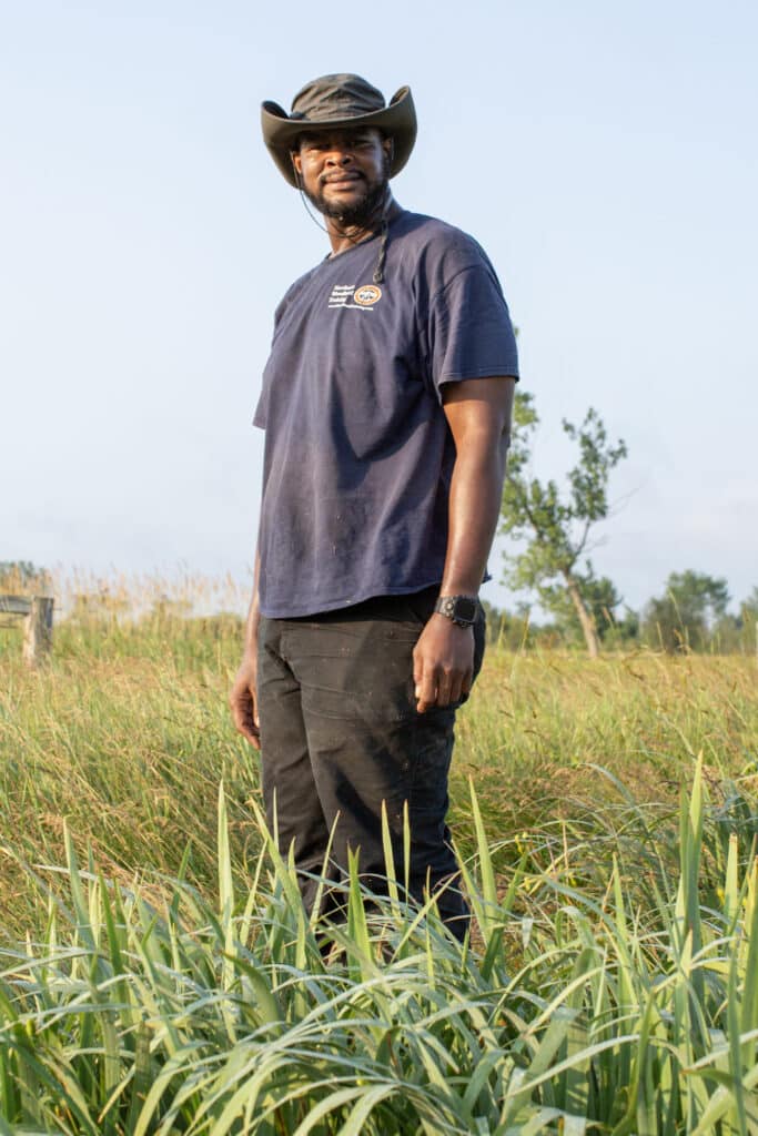 A man wearing a dark t-shirt, black pants, and a wide-brimmed hat stands in a grassy field under a clear sky, looking at the camera and smiling slightly.