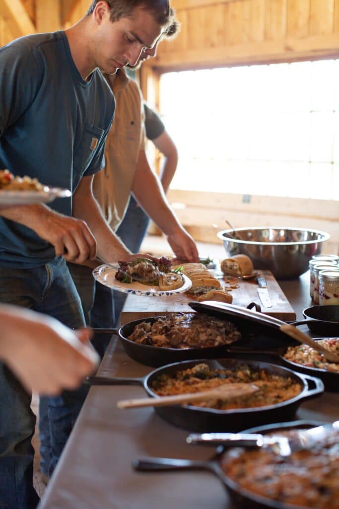 A man stands at a buffet table in a rustic setting, serving himself food from various dishes in cast iron pans. Other people are also helping themselves, and sunlight streams through large windows in the background.