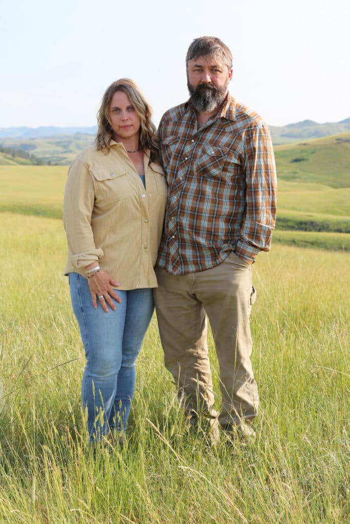 A woman and a man stand side by side in a grassy field with hills in the background, both wearing casual long-sleeve shirts and jeans, looking at the camera.