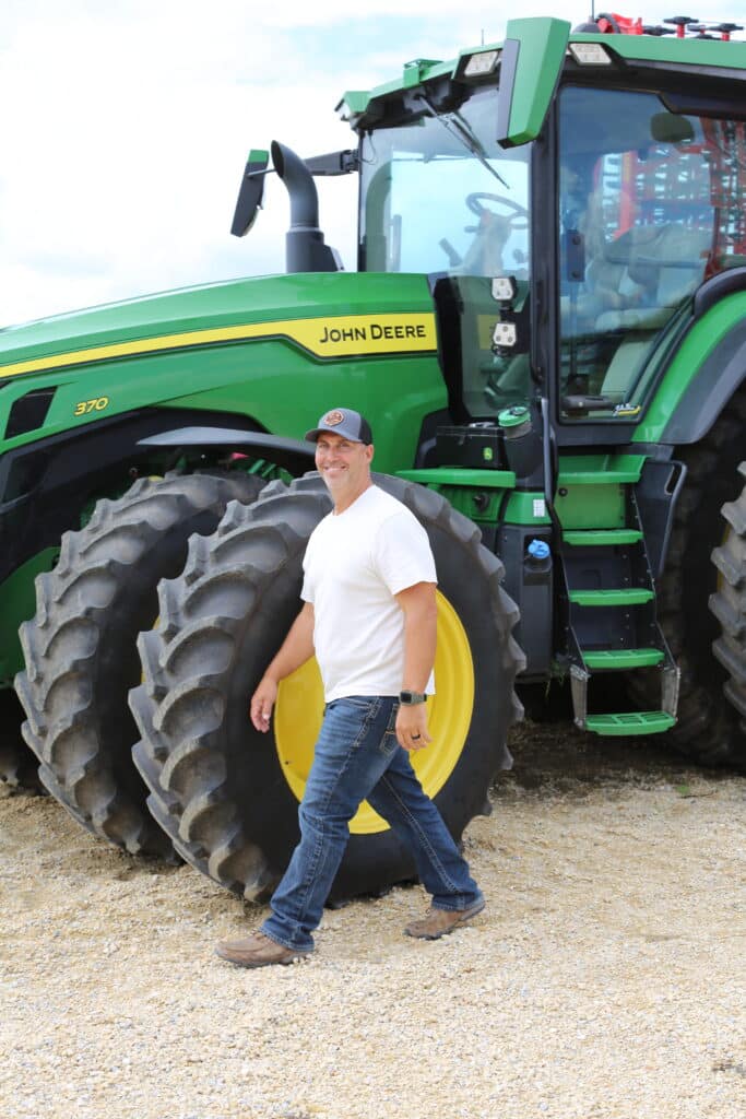 A man wearing a white t-shirt, jeans, and a cap walks in front of a large green John Deere tractor with four big tires on gravel.