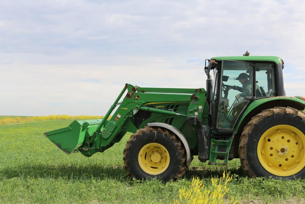 A person drives a green tractor with yellow wheels across a grassy field under a partly cloudy sky. The tractor's front loader is slightly raised.