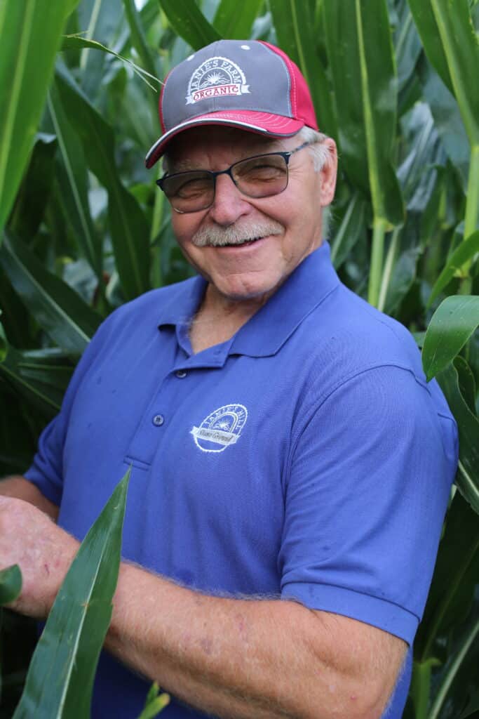 An older man wearing glasses, a red and gray cap, and a blue polo shirt smiles while standing among tall green corn plants in a field.
