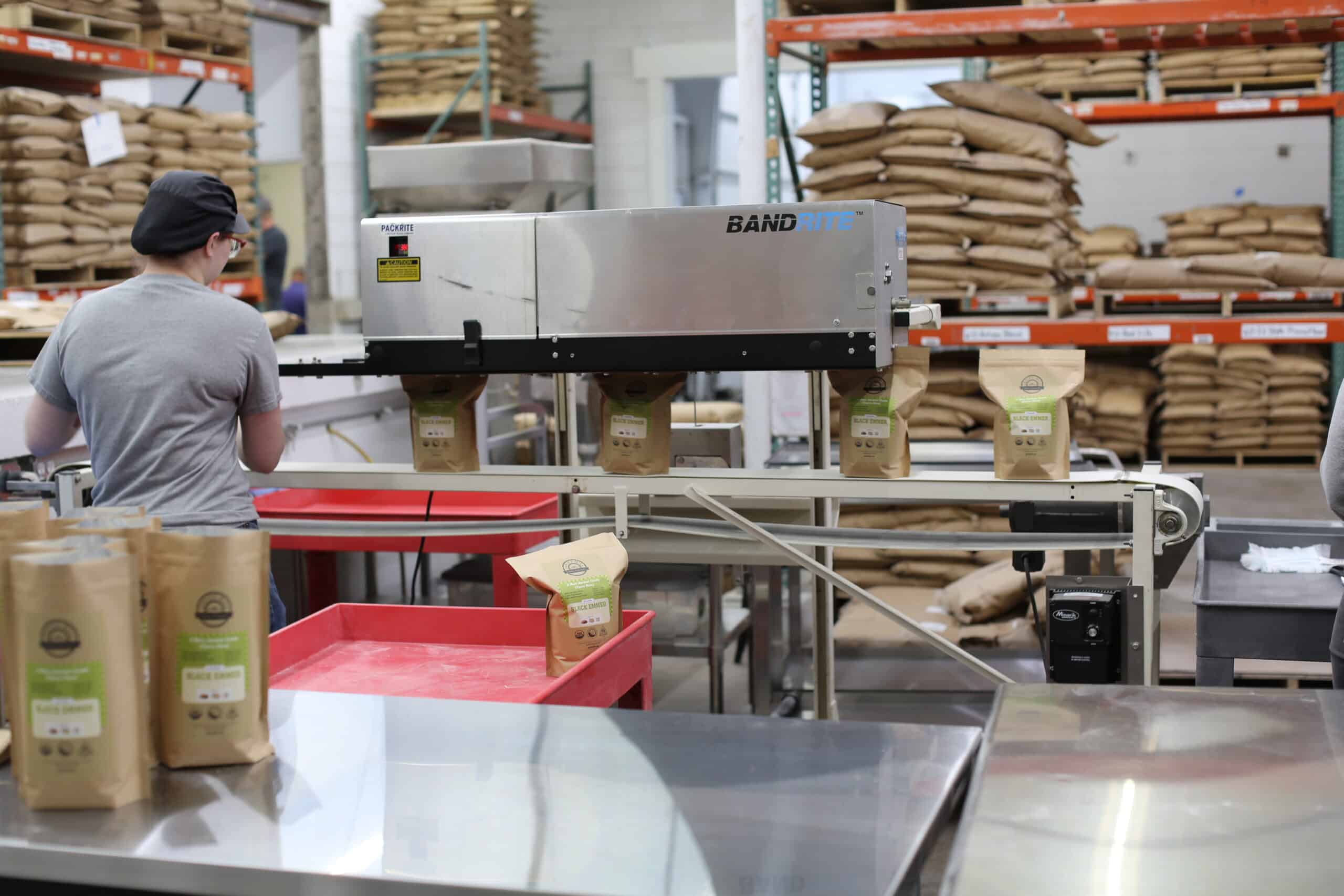 A person works at a packaging station in a factory, sealing bags of coffee on a conveyor belt. Shelves stacked with large brown sacks fill the background.