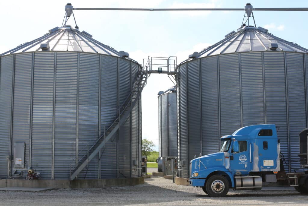 Two large metal grain silos with exterior staircases are shown side by side on a gravel lot. A blue semi-truck is parked in front of the silos, and the sky is bright with some clouds.