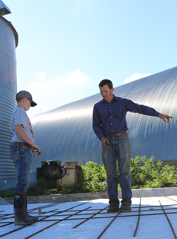 A man and a boy stand on a construction site with metal grids laid on the ground; the man is pointing and explaining something to the boy, with large farm buildings in the background under a blue sky.