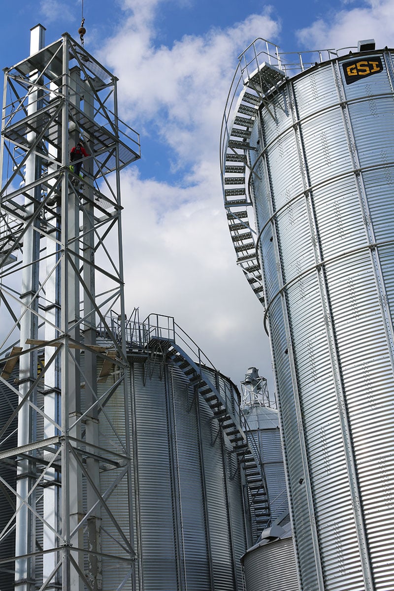 Silver metal grain silos and a tall metal structure stand against a partly cloudy blue sky. Stairs and platforms wrap around the silos, and a person in safety gear is climbing the metal tower.