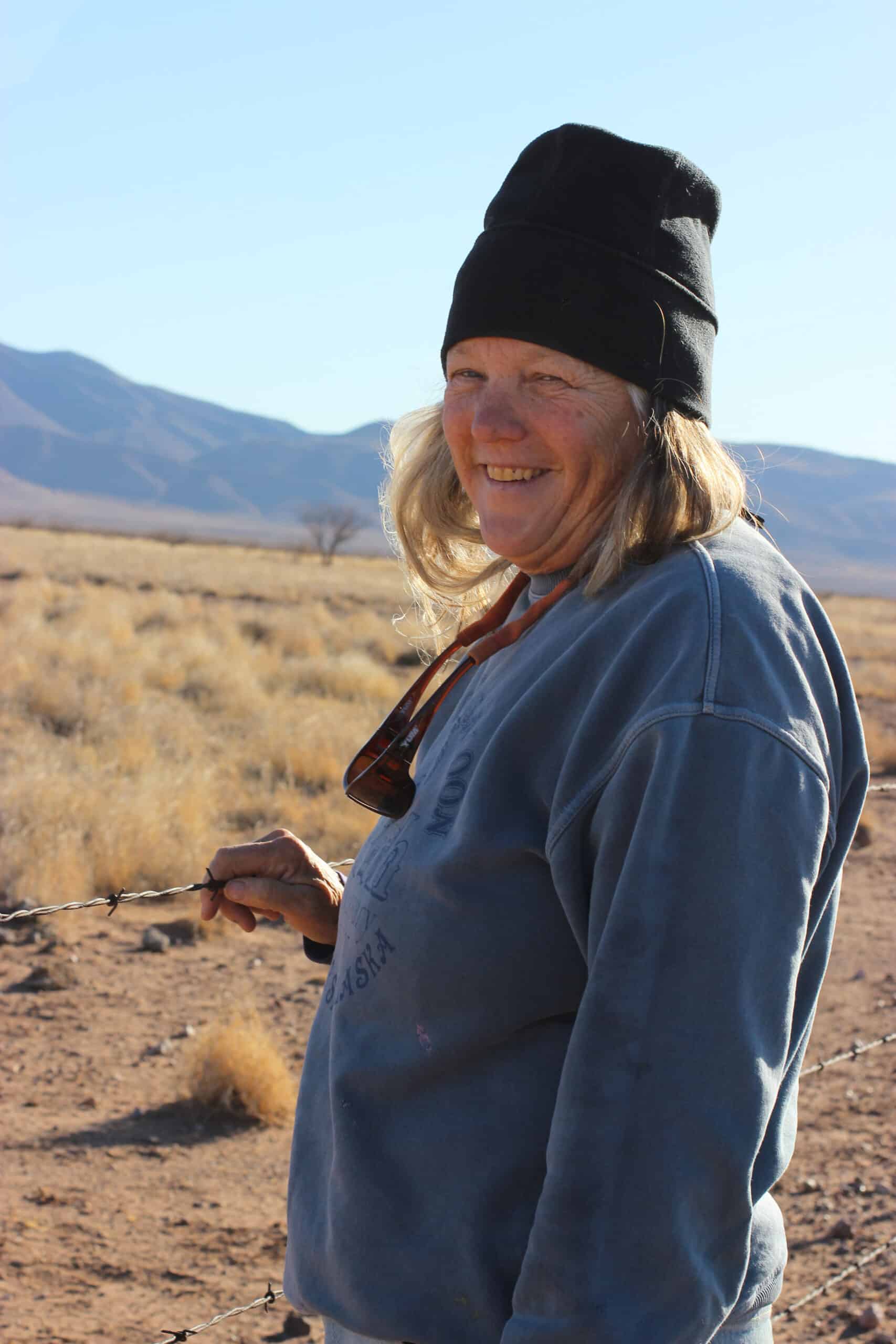 A person wearing a black beanie and blue sweatshirt smiles while standing in a dry, grassy landscape with distant mountains under a clear sky.