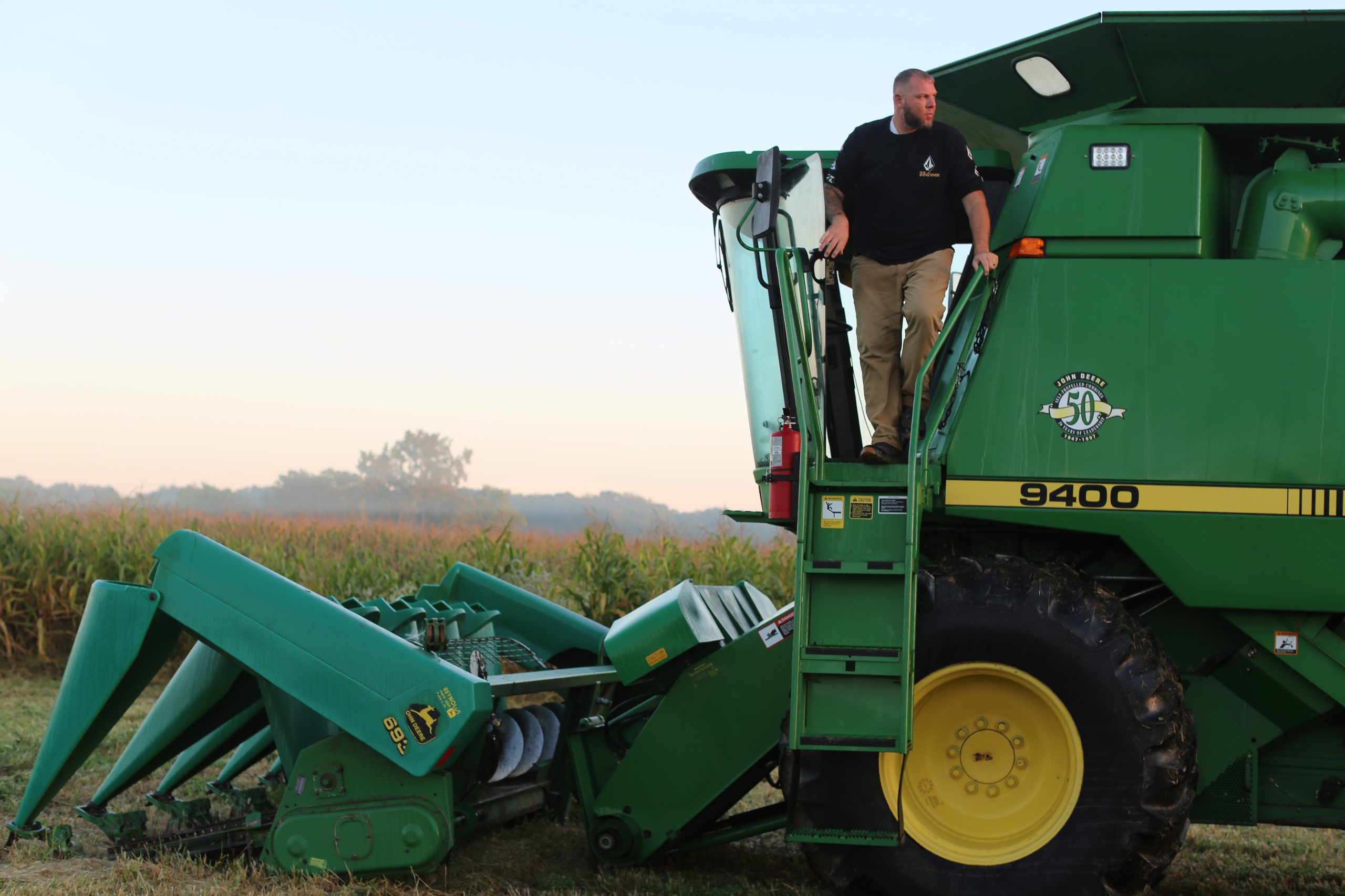 A farmer climbs down from the cab of a green John Deere 9400 combine harvester in a cornfield.
