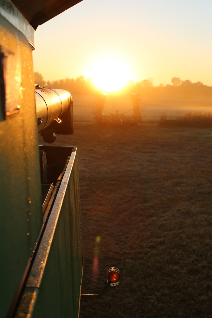The side of a green agricultural machine in a field at sunrise, with a piece of farm equipment visible in the distance.