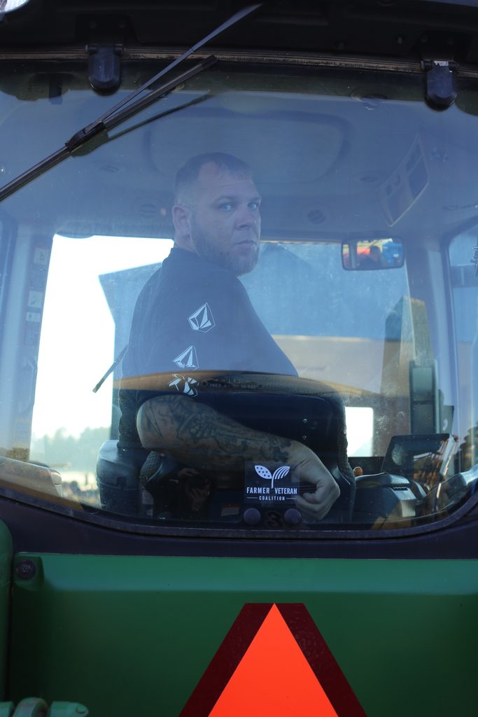 A man with a beard and tattoos sits in the cab of a tractor, holding a sign that reads “Farmer Veteran Coalition.”