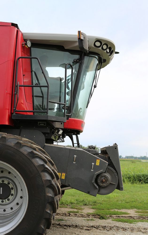 A close-up view of a red combine harvester parked on a grassy field, showing the cab with large windows, a metal ladder, and part of the front tire. The sky is cloudy in the background.